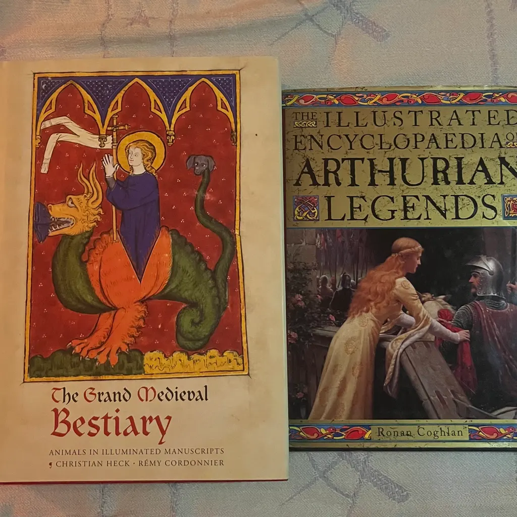 Two books: “The Grand Medieval Bestiary” with illuminated manuscript cover showing figure in blue robes within red arched frame, and “Illustrated Encyclopaedia of Arthurian Legends” with medieval scene cover.